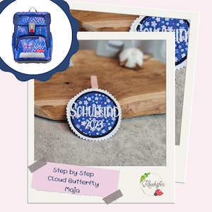 Könnte beinhalten: Ein blauer Rucksack mit einem Schmetterlingsdesign und dem Text "Step by Step Cloud Butterfly maja". Außerdem ist ein blauer und weißer runder Patch mit dem Text "Schulkind 2023" zu sehen.
