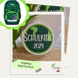 Könnte beinhalten: Grüner und schwarzer ergonomischer Rucksack mit Blitz-Akzenten und einem lila Design. Das Bild zeigt auch einen runden Aufnäher mit dem Text "SCHULKIND 2024" und einem grünen Blitz.