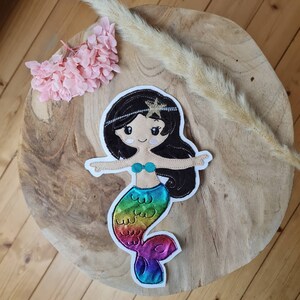 Könnte beinhalten: Eine Regenbogen-Meerjungfrau-Applikation mit schwarzen Haaren und einem goldenen Stern auf dem Kopf. Die Meerjungfrau lächelt und trägt einen türkisfarbenen Muschel-BH.