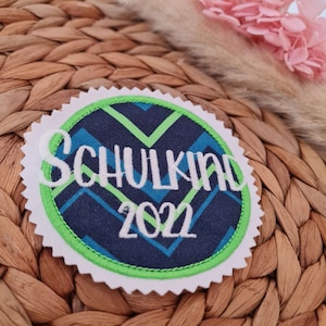 Könnte beinhalten: Ein weißer, grüner und blauer runder Patch mit einer weißen Stickerei, auf der "Schulkind 2022" steht.