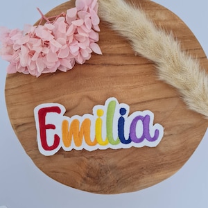 Könnte beinhalten: Ein gestickter Patch in Regenbogenfarben mit dem Namen "Emilia" auf weißem Hintergrund. Der Patch befindet sich auf einer Holzoberfläche.