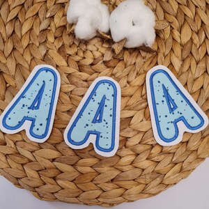 Könnte beinhalten: Drei blaue Filzbuchstaben-Patches mit weißen Punkten, jeder Buchstabe ist ein A, die Buchstaben befinden sich auf einem geflochtenen Weidenhintergrund.