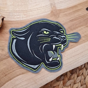 Könnte beinhalten: Schwarzer Pantherkopf-Stickerei-Aufnäher mit grünem Umriss und gelben Augen. Der Aufnäher befindet sich auf einer Holzoberfläche.