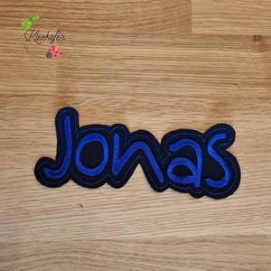 Könnte beinhalten: Ein blauer und schwarzer gestickter Aufnäher mit dem Namen "Jonas" in einer stilisierten Schrift.