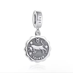 May include: A silver charm with a Taurus zodiac design. The charm features a round pendant with a scalloped edge, a bull illustration, stars, and the word "TAURUS". The charm has a loop for attaching to a bracelet or necklace.