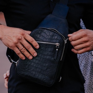 May include: A black leather crossbody bag with a crocodile-embossed design. The bag has a zipper closure and a silver strap.