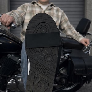 Może przedstawiać: Czarny but sportowy Puma z teksturowaną podeszwą i czarnym paskiem. Podeszwa ma logo Puma. But sportowy jest noszony z niebieskimi dżinsami i koszulą w kratę. W tle znajduje się motocykl.