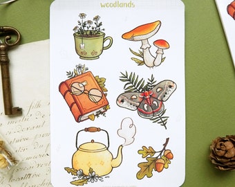 May include: A sheet of stickers with a woodland theme. The stickers feature a teapot, a book with glasses, a mushroom, a moth, a cup of tea with flowers, and an acorn branch. The stickers are on a white background with the word "woodlands" at the top.