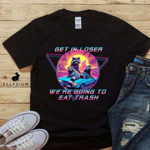 May include: Black t-shirt with a retro 80s design featuring three raccoons in a car with the text "Get in loser, we're going to eat trash".