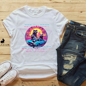 May include: White t-shirt with a graphic of raccoons in a car. The graphic is in a retro 80s style with bright colors and a pixelated look. The text on the shirt says "Get in Loser, We're Going to Eat Trash".