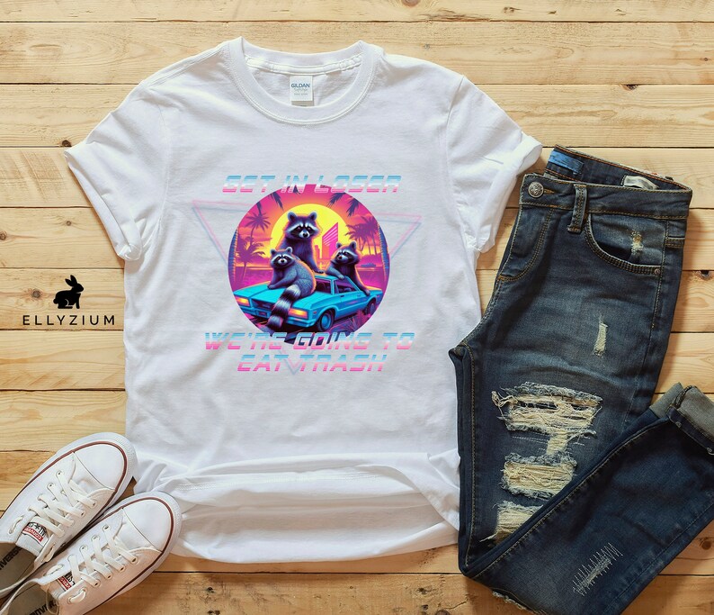 May include: White t-shirt with a graphic of raccoons in a car. The graphic is in a retro 80s style with bright colors and a pixelated look. The text on the shirt says "Get in Loser, We're Going to Eat Trash".