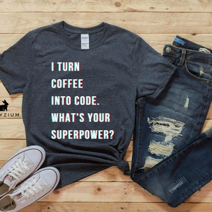 K&ouml;nnte beinhalten: Ein dunkelgraues T-Shirt mit dem Text "I turn coffee into code. What's your superpower?" in Glitch-Effekt. Das Shirt liegt auf einer Holzoberfl&auml;che mit einem Paar wei&szlig;en Sneakers und einer blauen Jeans.