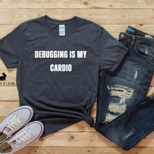 May include: A dark gray t-shirt with the text "Debugging is my cardio" in white with a glitch effect. The shirt is laid out on a wood surface with a pair of white sneakers and blue jeans.