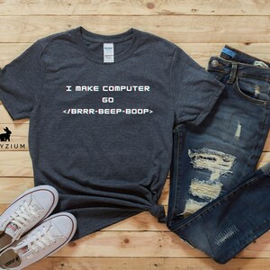 May include: A dark grey t-shirt with the text "I MAKE COMPUTER GO </BRRR-BEEP-BOOP>" printed on it. The shirt is laid out on a wooden surface with a pair of white trainers and blue jeans.