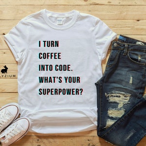 K&ouml;nnte beinhalten: Wei&szlig;es T-Shirt mit dem Text "I turn coffee into code. What's your superpower?" darauf gedruckt. Das Shirt liegt auf einer Holzoberfl&auml;che mit einer blauen Jeans und einem Paar wei&szlig;en Turnschuhen.