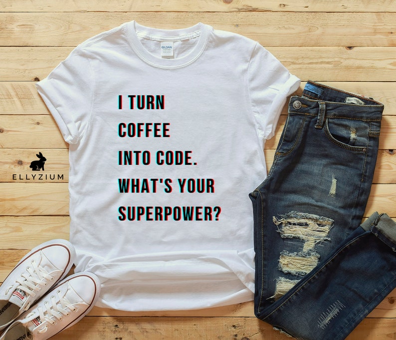 K&ouml;nnte beinhalten: Wei&szlig;es T-Shirt mit dem Text "I turn coffee into code. What's your superpower?" darauf gedruckt. Das Shirt liegt auf einer Holzoberfl&auml;che mit einer blauen Jeans und einem Paar wei&szlig;en Turnschuhen.