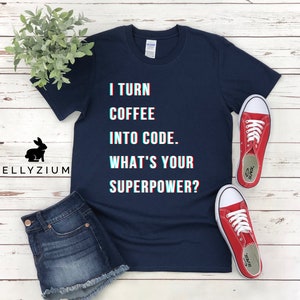 K&ouml;nnte beinhalten: Ein marineblaues T-Shirt mit dem Text "I turn coffee into code. What's your superpower?" in einer Glitch-Schriftart. Das Shirt liegt flach auf einer wei&szlig;en Holzoberfl&auml;che mit einem Paar roten Turnschuhen und einer Jeans-Shorts.