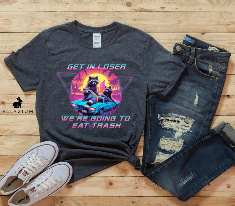 May include: A dark gray t-shirt with a retro-style graphic of raccoons in a car. The graphic includes the text "Get in Loser, We're Going to Eat Trash".