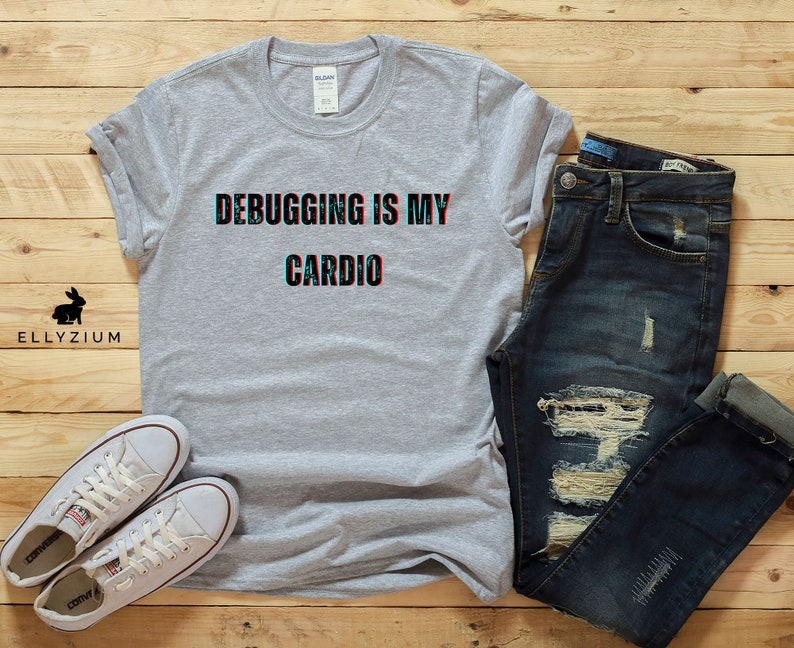 May include: A gray t-shirt with the text "Debugging is my cardio" in a glitch effect. The shirt is laid out on a wooden surface with a pair of white sneakers and blue jeans.