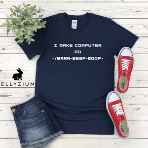 May include: A navy blue t-shirt with the text "I MAKE COMPUTER GO <BRRR-BEEP-BOOP>" printed on the front. The shirt is laid flat on a white wooden surface.