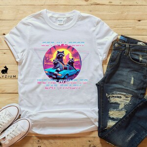 May include: White t-shirt with a graphic of raccoons in a car. The graphic is in a retro 80s style with bright colors and a pixelated look. The text on the shirt says "Get in Loser, We're Going to Eat Trash".