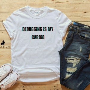 May include: White t-shirt with the text "Debugging is my cardio" in a glitched font. The shirt is laid out on a wooden surface with a pair of blue jeans and a pair of white sneakers.