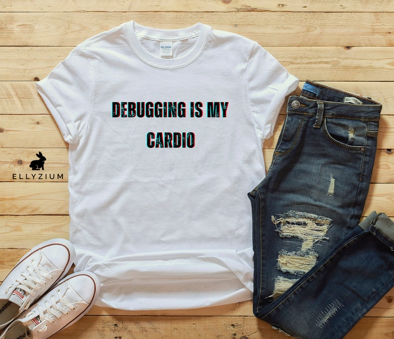 May include: White t-shirt with the text "Debugging is my cardio" in a glitched font. The shirt is laid out on a wooden surface with a pair of blue jeans and a pair of white sneakers.