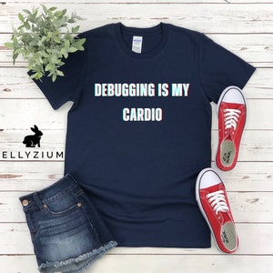 May include: A navy blue t-shirt with the text "Debugging is my cardio" in white with a glitch effect. The shirt is laid flat on a white wooden surface with a pair of red sneakers.