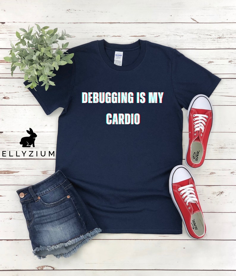 May include: A navy blue t-shirt with the text "Debugging is my cardio" in white with a glitch effect. The shirt is laid flat on a white wooden surface with a pair of red sneakers.