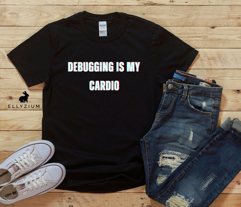 May include: Black t-shirt with the text "Debugging is my cardio" in white with a glitch effect. The shirt is laid flat on a wooden surface with a pair of white sneakers and blue jeans.