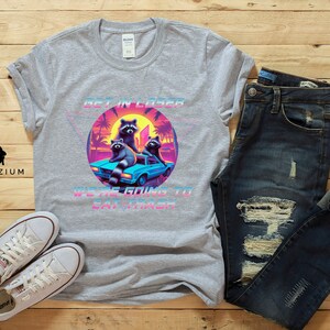 May include: A gray t-shirt with a retro-style graphic of raccoons in a car. The graphic says "Get in Loser, We're Going to Eat Trash".