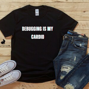 May include: Black t-shirt with the text "Debugging is my cardio" in white with a 3D effect. The shirt is laid flat on a wooden surface with a pair of white sneakers and blue jeans.