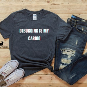 May include: A dark gray t-shirt with the text "Debugging is my cardio" in white with a glitch effect. The shirt is laid out on a wood surface with a pair of white sneakers and blue jeans.