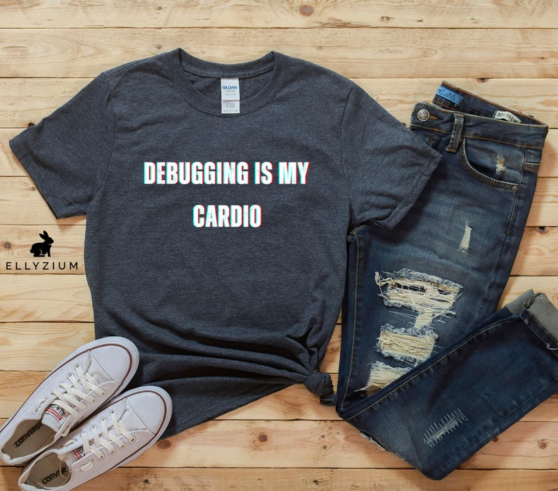 May include: A dark gray t-shirt with the text "Debugging is my cardio" in white with a glitch effect. The shirt is laid out on a wood surface with a pair of white sneakers and blue jeans.
