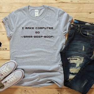 May include: A grey t-shirt with the text "I MAKE COMPUTER GO </BRRR-BEEP-BOOP>" printed on it. The shirt is paired with blue jeans and white trainers.