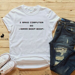 May include: White t-shirt with the text "I make computer go <brr-beep-boop>" printed on it. The shirt is laid out on a wooden surface with a pair of blue jeans and white trainers.