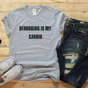 May include: A gray t-shirt with the text "Debugging is my cardio" in a glitch effect. The shirt is laid out on a wooden surface with a pair of white sneakers and blue jeans.