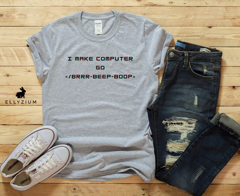 May include: A grey t-shirt with the text "I MAKE COMPUTER GO </BRRR-BEEP-BOOP>" printed on it. The shirt is paired with blue jeans and white trainers.
