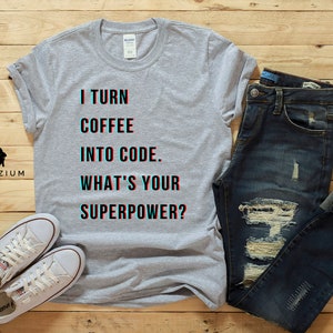 K&ouml;nnte beinhalten: Ein graues T-Shirt mit dem Text "I turn coffee into code. What's your superpower?" in Schwarz und Blau. Das Shirt wird mit einer blauen Jeans und wei&szlig;en Turnschuhen kombiniert.