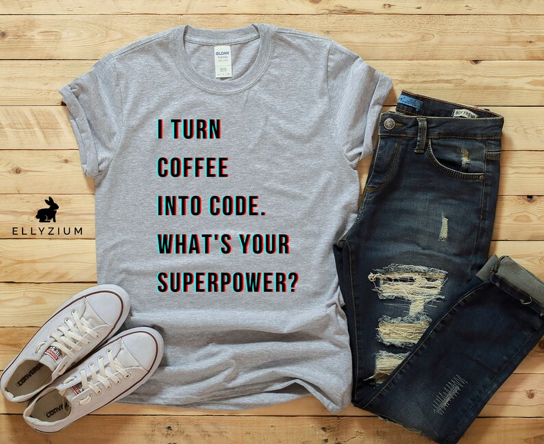 K&ouml;nnte beinhalten: Ein graues T-Shirt mit dem Text "I turn coffee into code. What's your superpower?" in Schwarz und Blau. Das Shirt wird mit einer blauen Jeans und wei&szlig;en Turnschuhen kombiniert.
