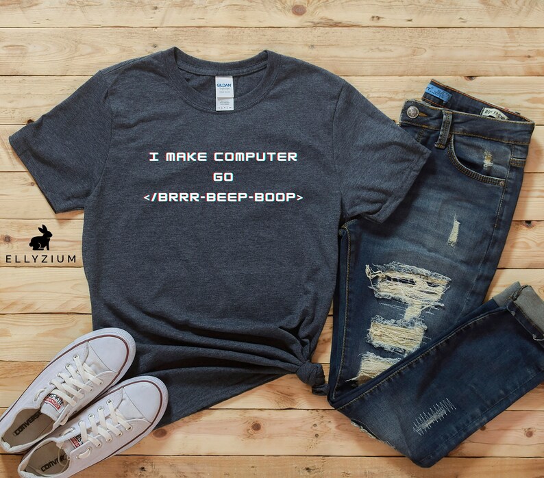 May include: A dark grey t-shirt with the text "I MAKE COMPUTER GO </BRRR-BEEP-BOOP>" printed on it. The shirt is laid out on a wooden surface with a pair of white trainers and blue jeans.