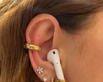 May include: An ear featuring various pieces of jewellery. A gold ear cuff with small details is at the top. A white earbud is held in place by a gold ear wrap. Other earrings include a flower-shaped stud and a small hoop.