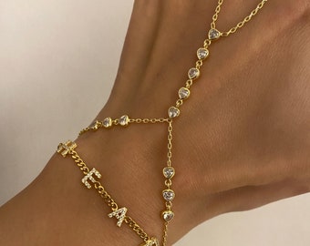 May include: A gold-colored hand chain with the letters "FAIR" spelled out in small, clear crystals. The chain features heart-shaped crystal accents and connects to a ring. The jewelry is worn on a hand.