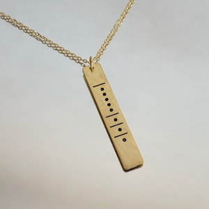 May include: A gold necklace with a rectangular pendant featuring a vertical line and a series of dots, resembling a code or a pattern.