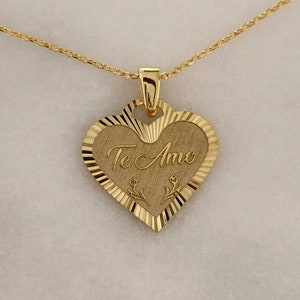 May include: A gold heart-shaped pendant necklace with the words "Te Amo" engraved on it. The pendant has a textured edge and is attached to a delicate gold chain.