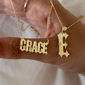 May include: Two gold necklaces with name pendants. One necklace has the name "GRACE" in a stylised font. The other necklace has a single letter "E" pendant.