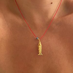 Op de afbeelding: Een gouden vishanger ketting aan een rode koord. De vishanger heeft gedetailleerde schubben en vinnen. Een kleine blauwe en witte kraal zit ook op het koord. De ketting wordt om de nek gedragen.