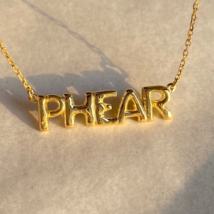 Könnte beinhalten: Eine goldene Kettenkette mit einem Namensschild-Anhänger, auf dem "PHEAR" in fetten Blockbuchstaben steht.