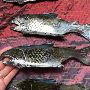 May include: Three elongated ceramic dishes shaped like fish, featuring a metallic, iridescent glaze with dark blue, green, and silver tones. Each dish has a textured scale pattern on the body, a defined head with an open mouth, and a finned tail.