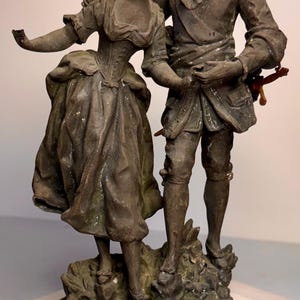 May include: A bronze-toned metal sculpture depicting two figures standing together on a base. The base features a dark green marble-like finish and a small rectangular plaque with the text "PASSAGE DU GUE" and "Par Dancoulet".